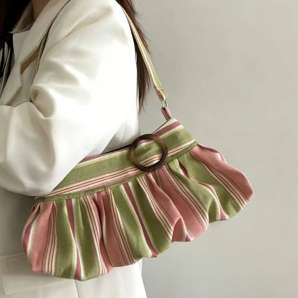 A person wearing a white blazer carries a pleated shoulder bag with pink and sage green stripes and a round brown ring detail on a light gray background.