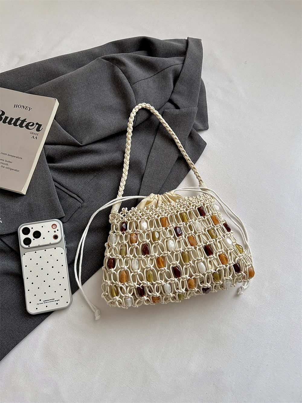 A cream macrame handbag with woven brown, amber, and white stone accents, styled on a light gray fabric surface next to a gray blazer, a white smartphone case, and a book.