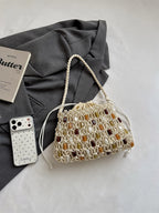 A cream macrame handbag with woven brown, amber, and white stone accents, styled on a light gray fabric surface next to a gray blazer, a white smartphone case, and a book.