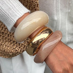 Close-up of a hand wearing two bangles, one beige and one gold, on a neutral background.