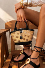 A woven straw and black leather bucket handbag with a top handle, held by a model sitting on a wooden bench. The model is also wearing black lace-up thong sandals with gold-tone hardware.