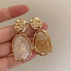 Gold earrings with stone-like designs held by a hand against a neutral background
