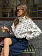 Woman sitting on a bench wearing a polka dot blouse and sunglasses, holding a drink.