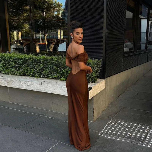 A woman wearing a brown off-the-shoulder maxi dress with a twist back design, standing on the sidewalk.
